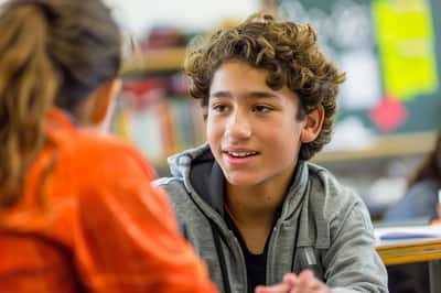 Zwei Mittelschüler unterhalten sich lachend an Schultischen in einem Klassenzimmer, was auf ein freundliches Gespräch hindeutet.